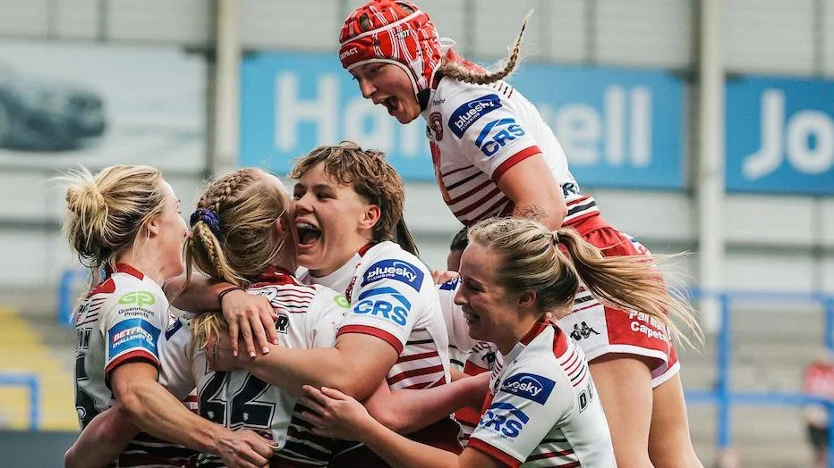 Wigan Warriors women players, in white kit with red trim,  surround try-scorer Ruby Hunter, with one jumping on the shoulders of a team-mate